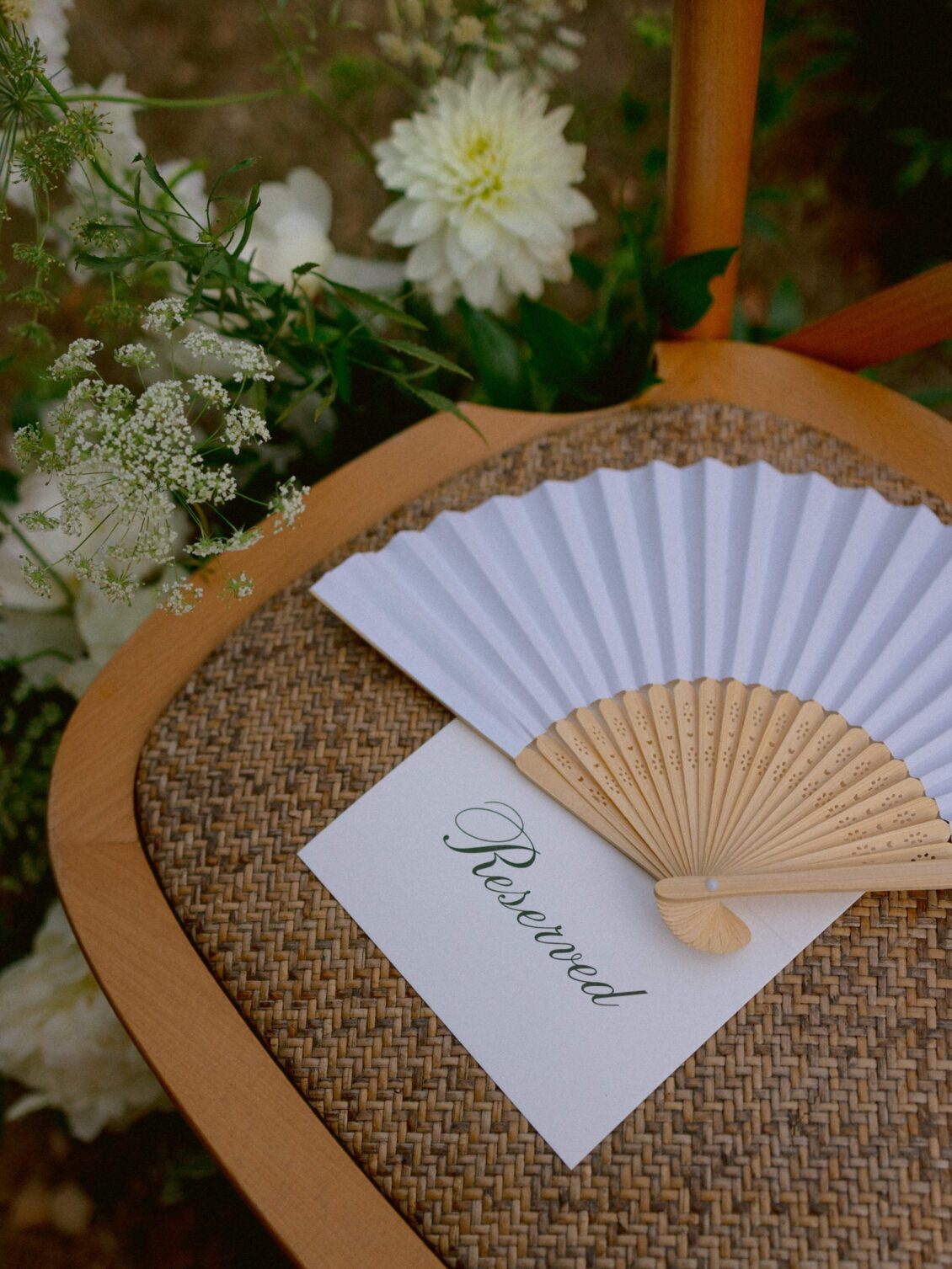 A white paper fan and a Reserved card resting on a rattan chair seat beside white flowers