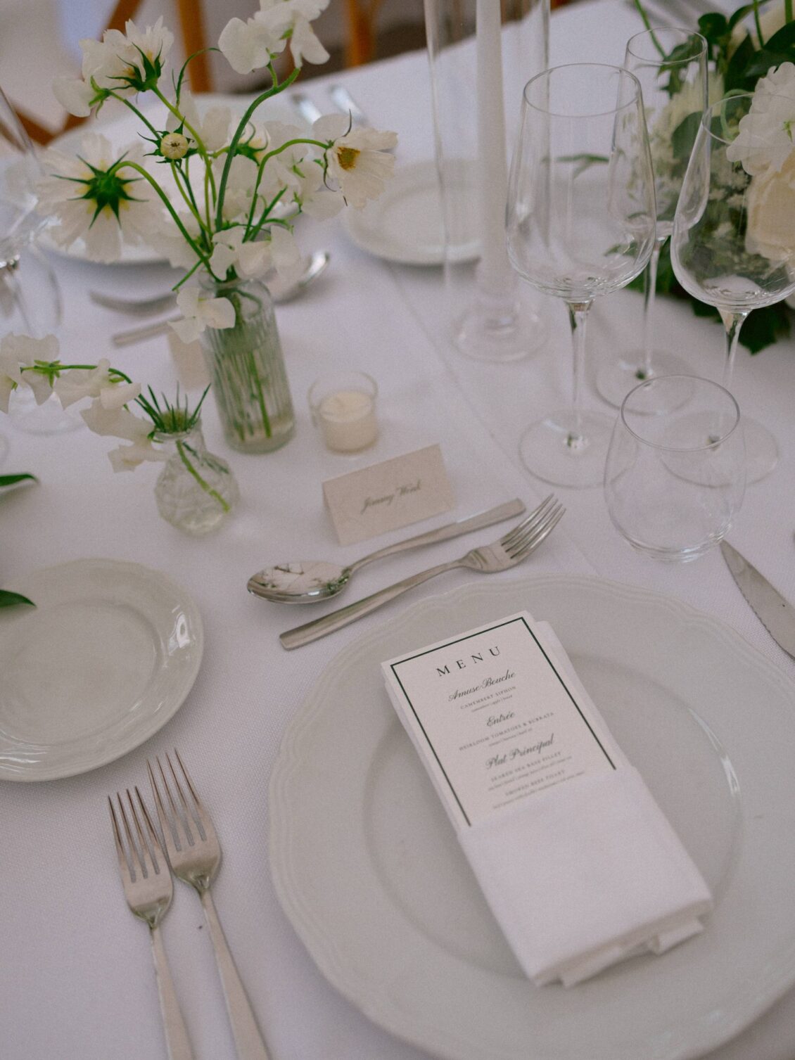 A white place setting with a printed menu, crystal glassware and bud vases of white flowers