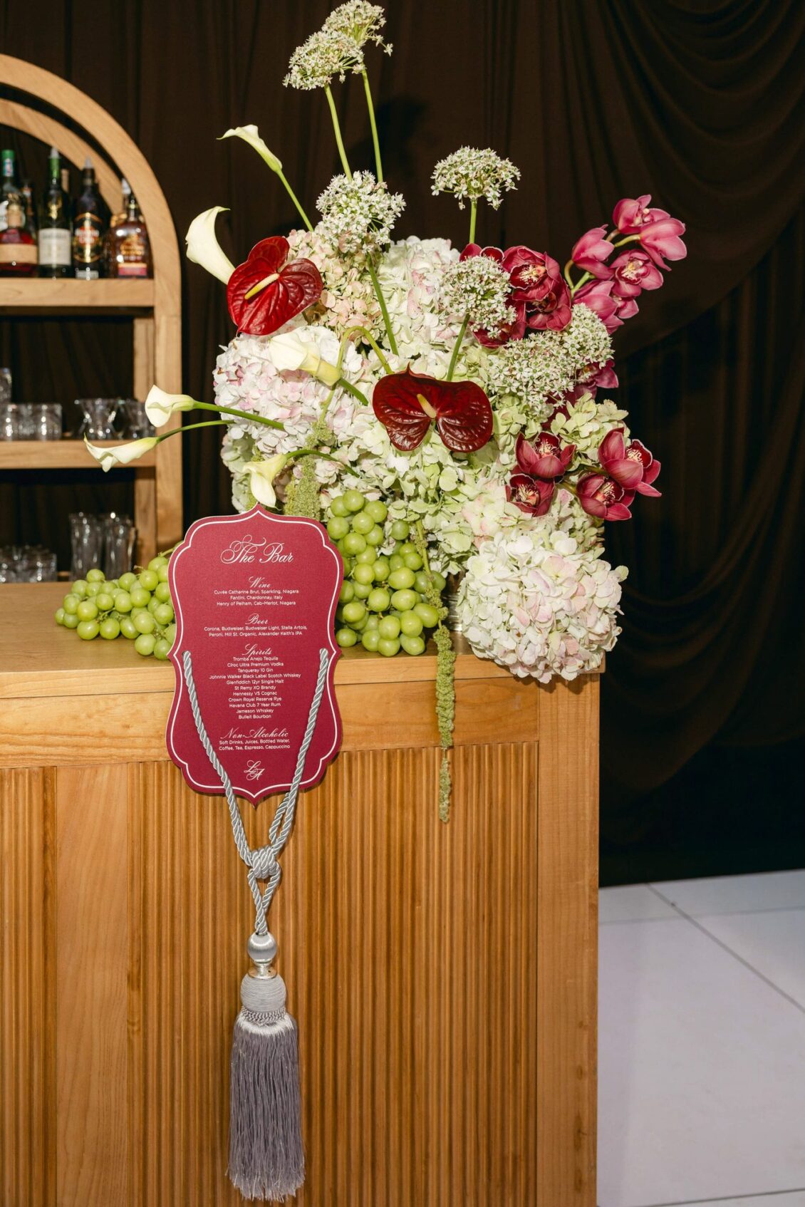 Bar arrangement of red anthuriums, white calla lilies, hydrangeas and grapes beside a burgundy menu with a tassel