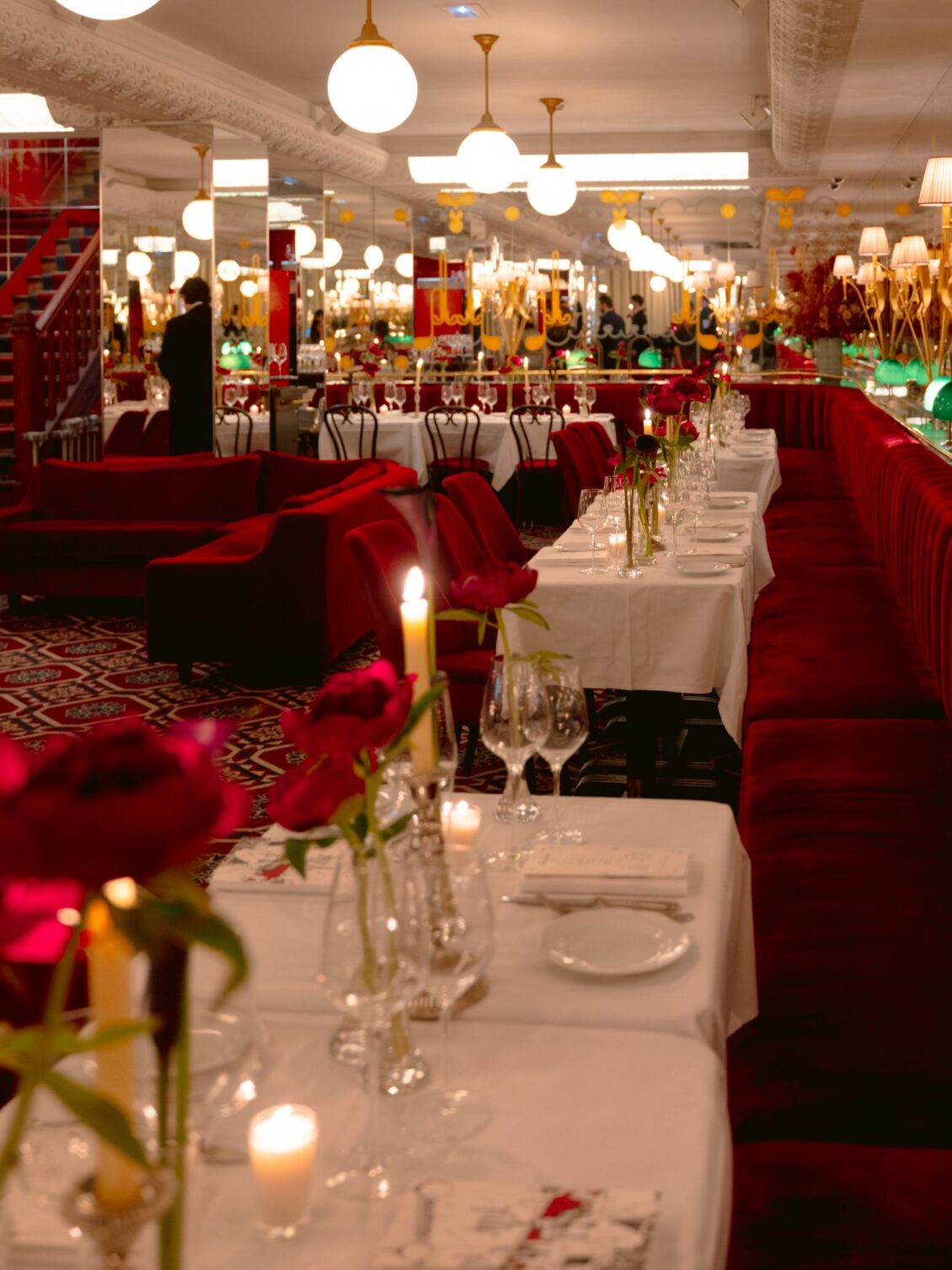 A long red velvet banquette lined with white-clothed tables, candles and red peonies at Thoumieux in Paris