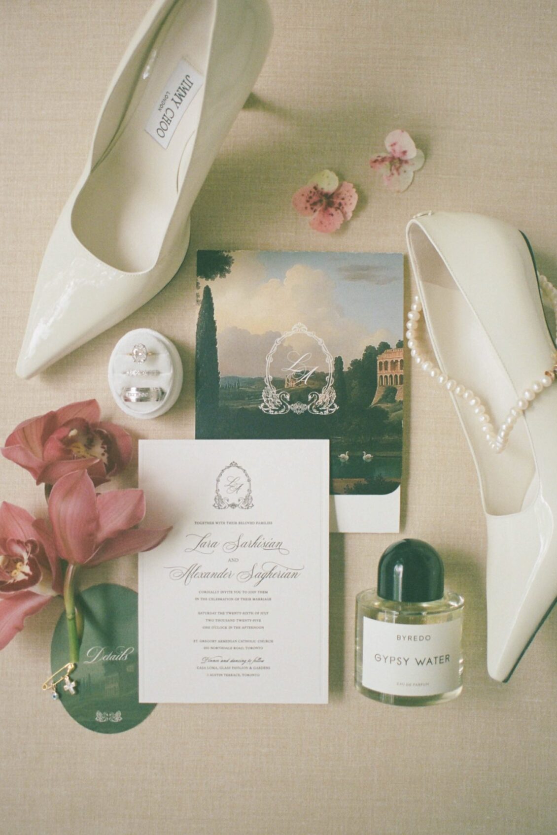 Wedding invitation with a swan crest beside a watercolour card, ivory pointed pumps, a perfume bottle and pink orchids