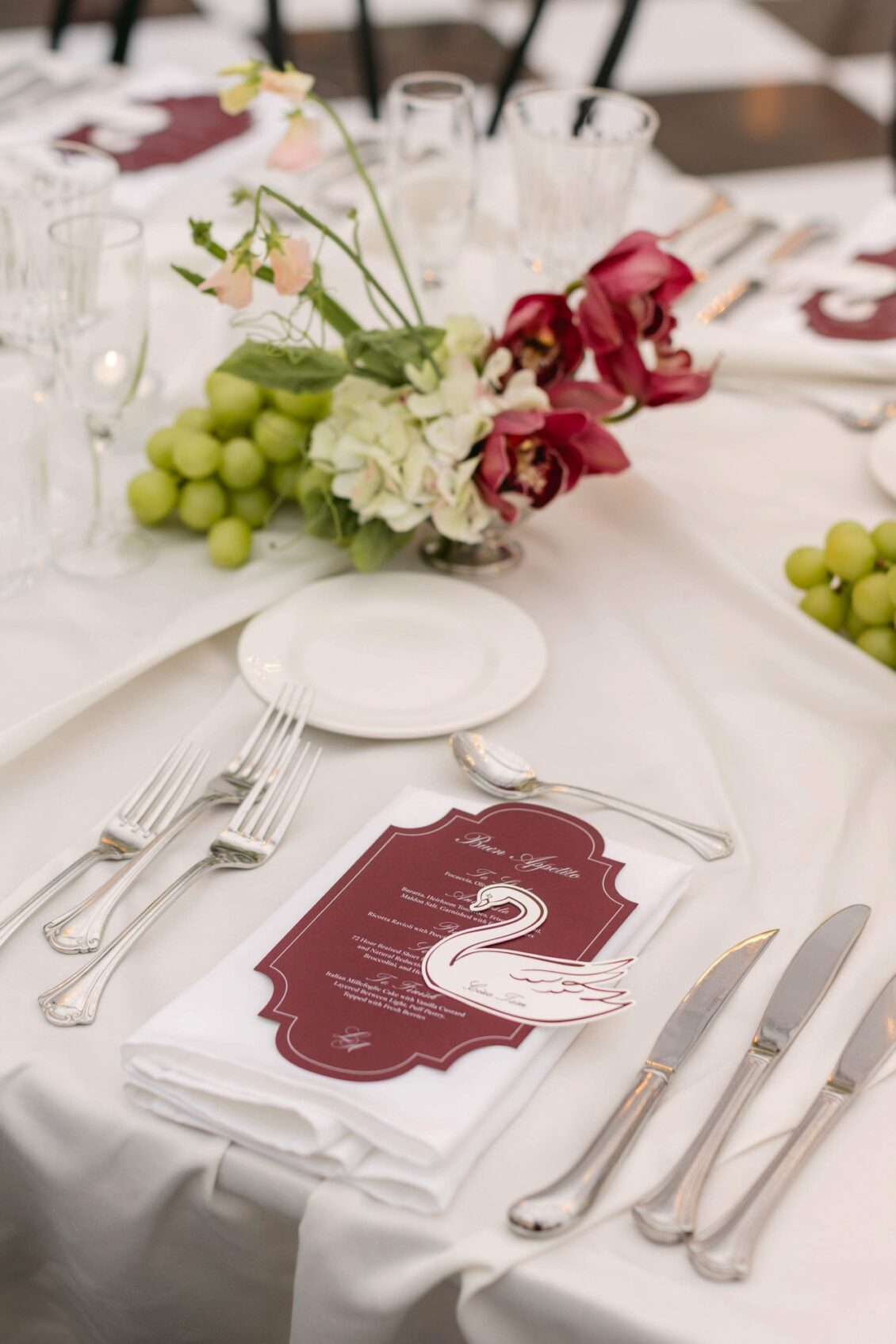 Place setting with a burgundy menu featuring a swan cut-out beside green grapes, red anthuriums and white hydrangea
