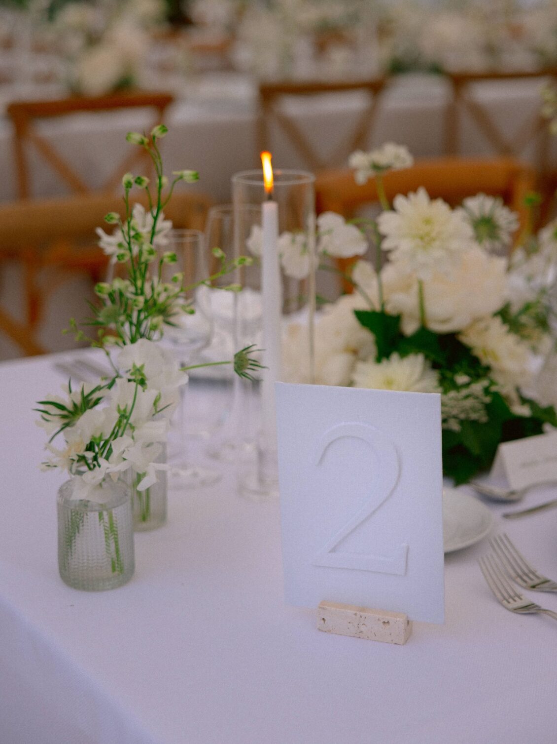 A frosted table number card beside white flowers in glass vases and a taper candle