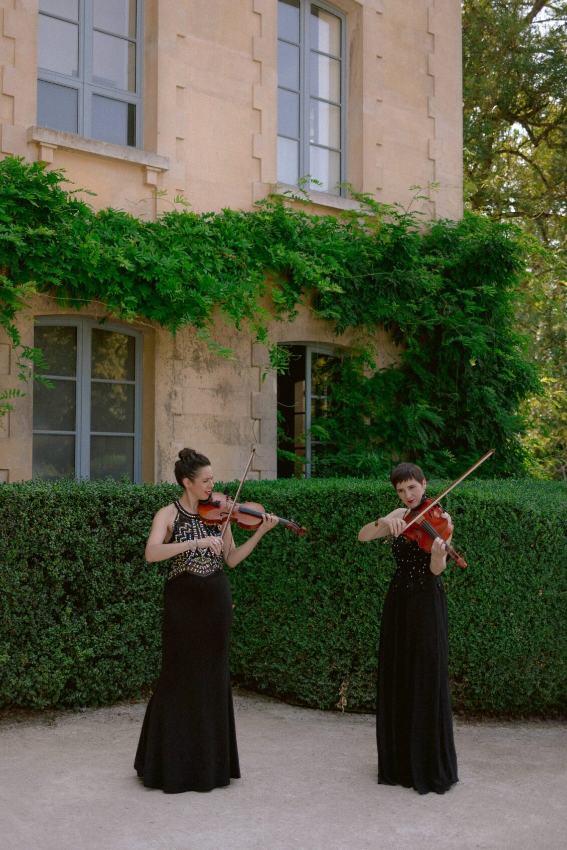 Two violinists in black gowns play in front of an ivy-covered château wall