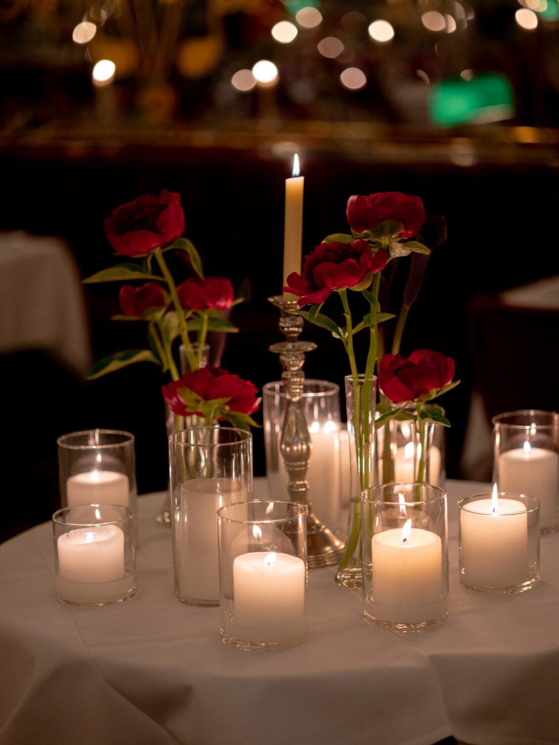 A round table centred with clustered votive candles, a silver candlestick and red peonies