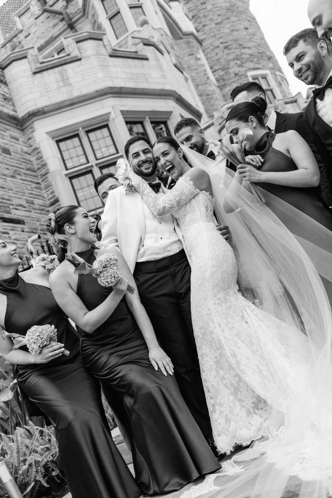 Black and white photo of the wedding party laughing around Lara and Alex in front of the castle