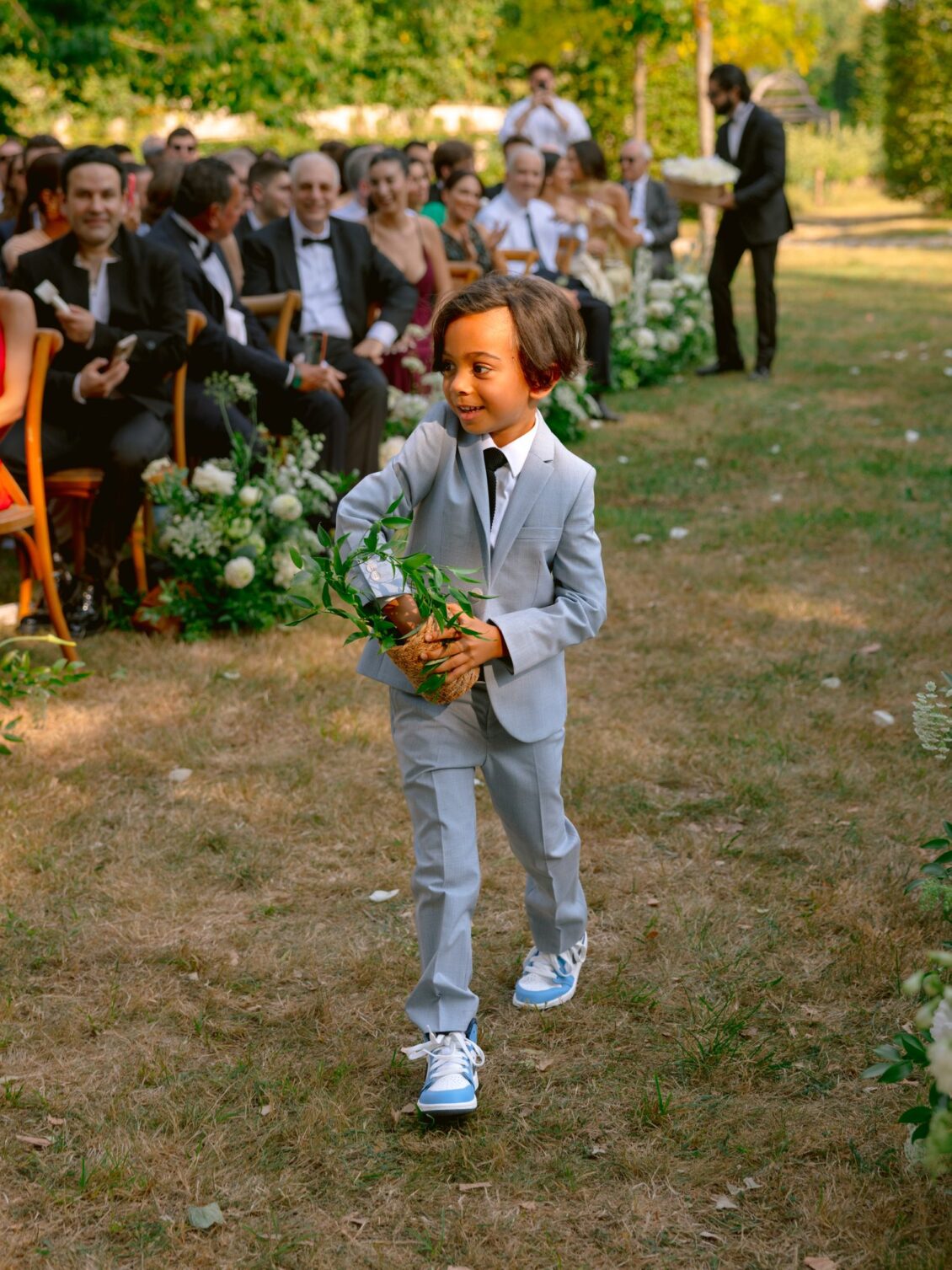 A smiling young boy in a pale grey suit carries a woven basket of greenery down the grass aisle as guests look on